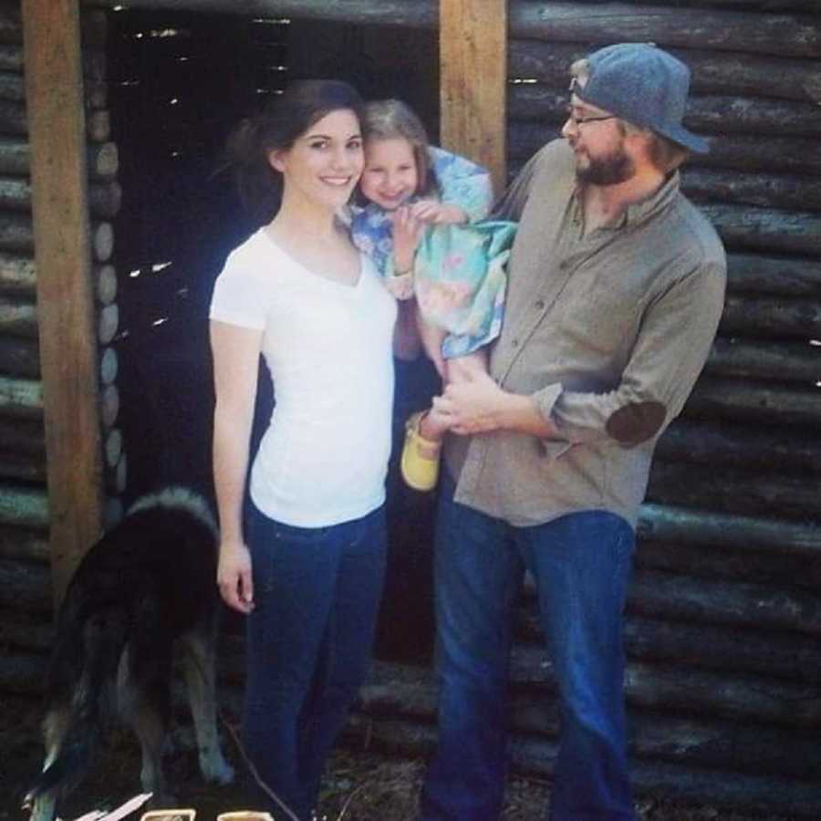 Husband and wife stand smiling outside log structure holding their daughter