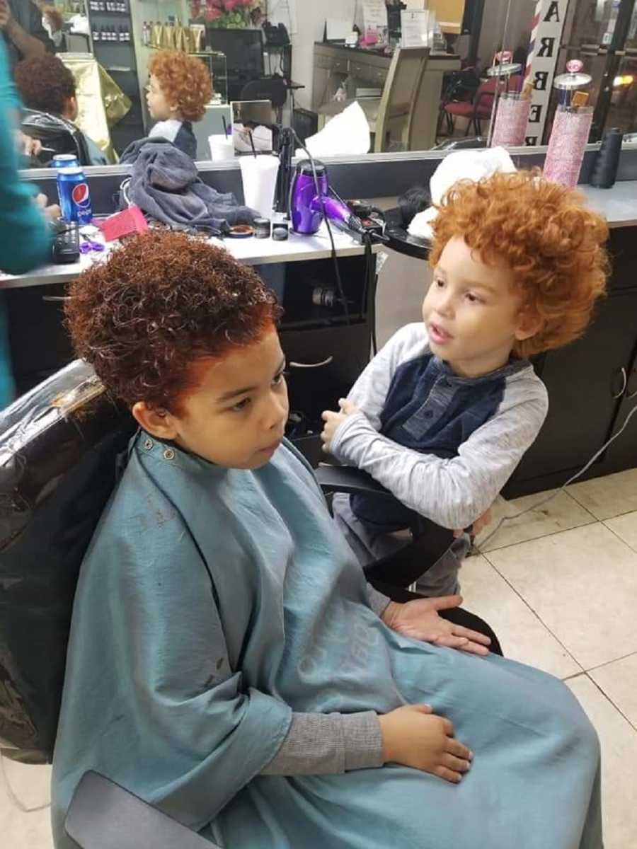 African American boy with red hair sits beside brother in barber shop chair who is dying is hair red