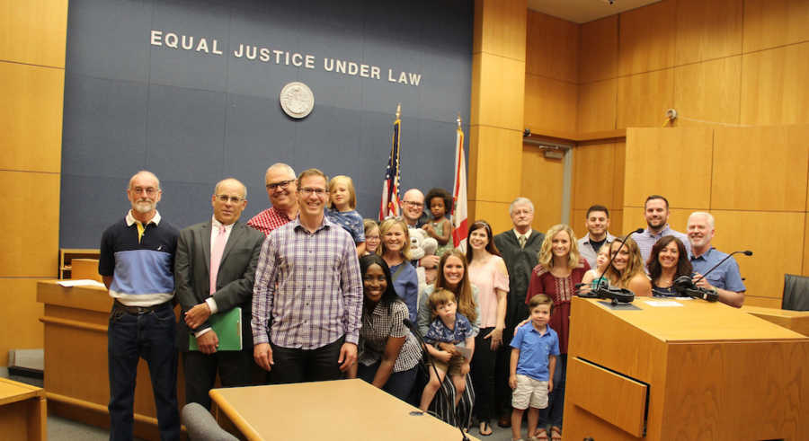 Husband and wife stand in adoption court beside their adopted daughter, three biological kids and family and friends