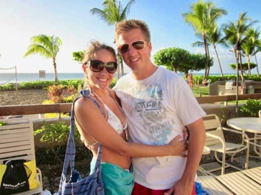 Woman who had postpartum depression stands smiling in swimsuit top with arms around husband with beach in background
