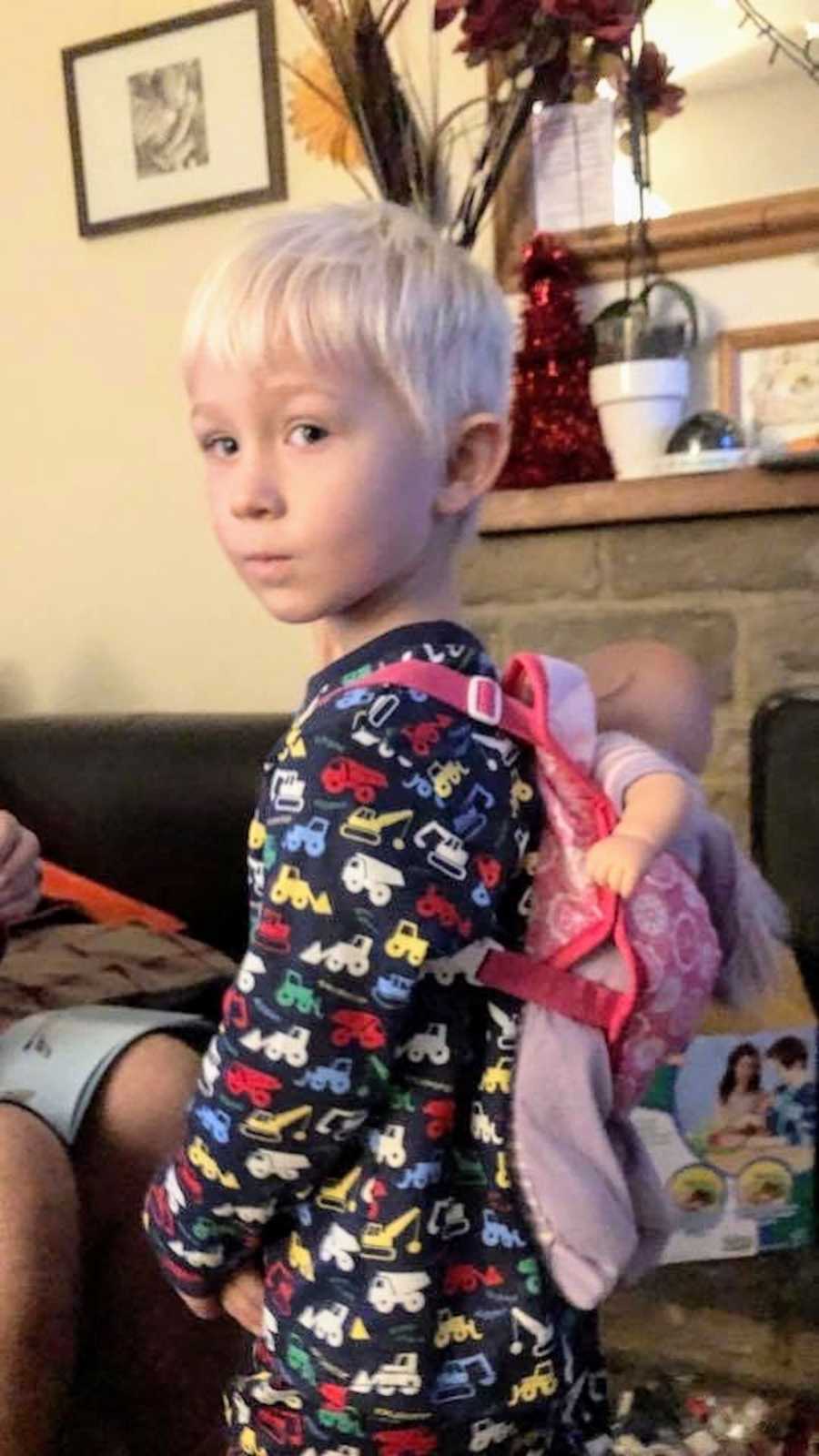 Little boy who still needs to be cuddled to sleep stands in home in pj's with pink backpack on with baby inside it