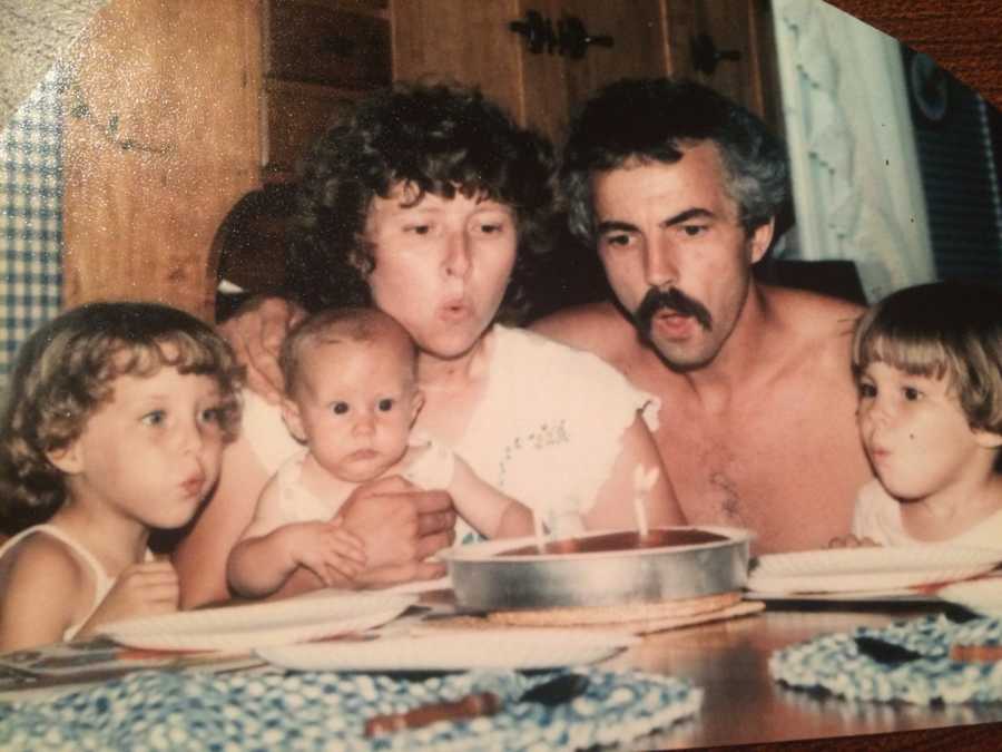 Mother and father sit at table with three young kids as they all blow out candles in cake