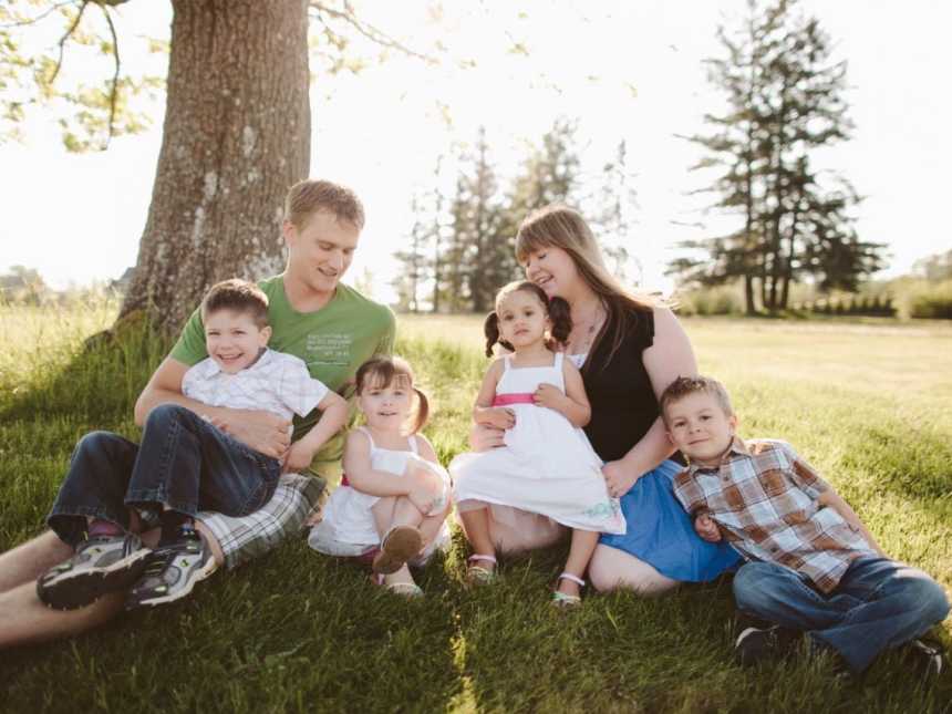 Husband and wife sit on ground outside beside tree with their four foster kids beside them