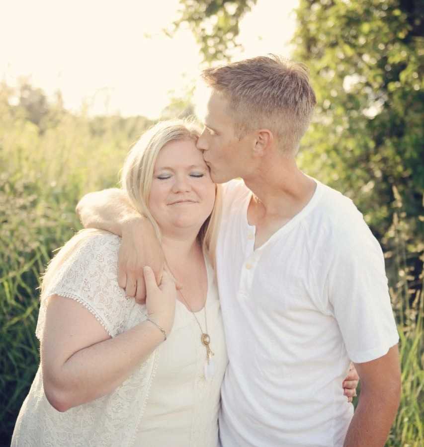 Husband stands with arms around wife kissing forehead of wife