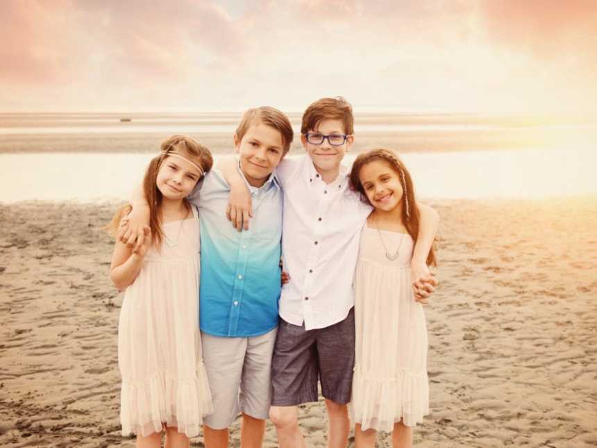 Four children in same foster home stand arm in arm on beach smiling
