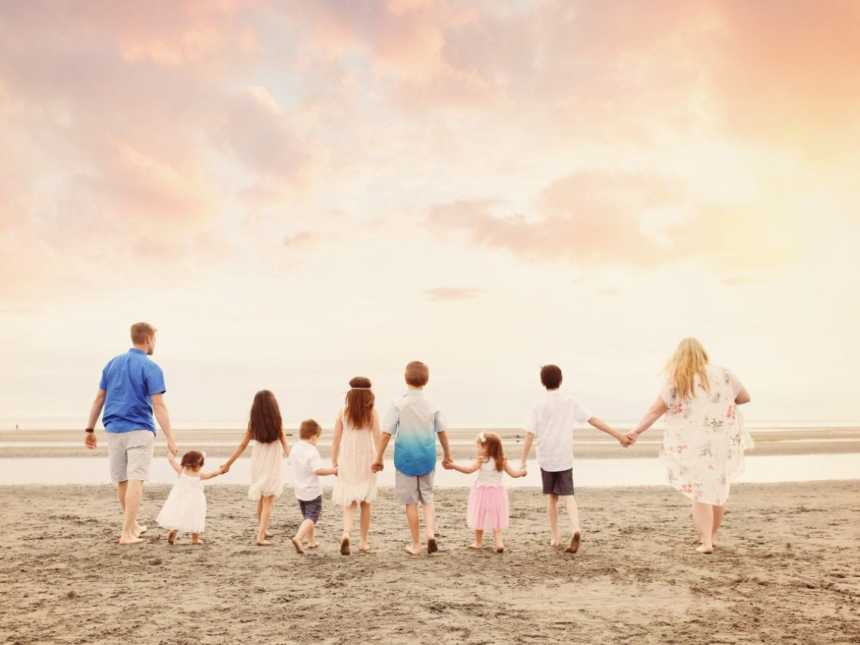 Husband and wife stands on beach holding hands with their seven foster children