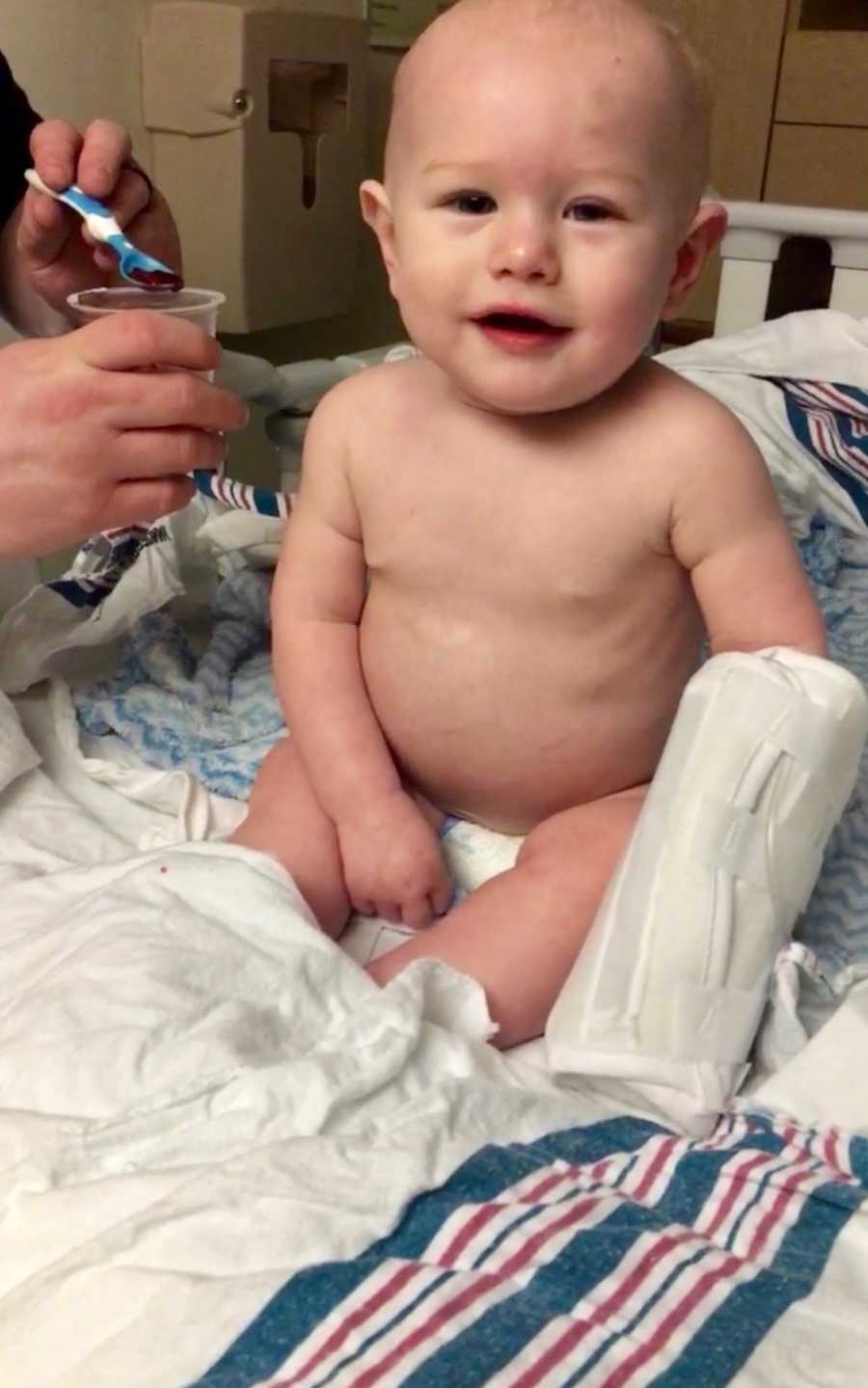 Baby with Transverse Myelitis sits up in hospital bed as someone holds up spoonful of Jell-O near his face