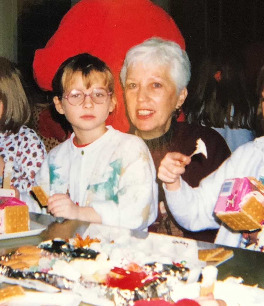 Little girl sits at table making gingerbread house with her grandmother smiling behind her