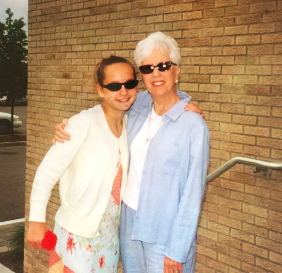 Teen stands smiling beside grandma in front of brick wall