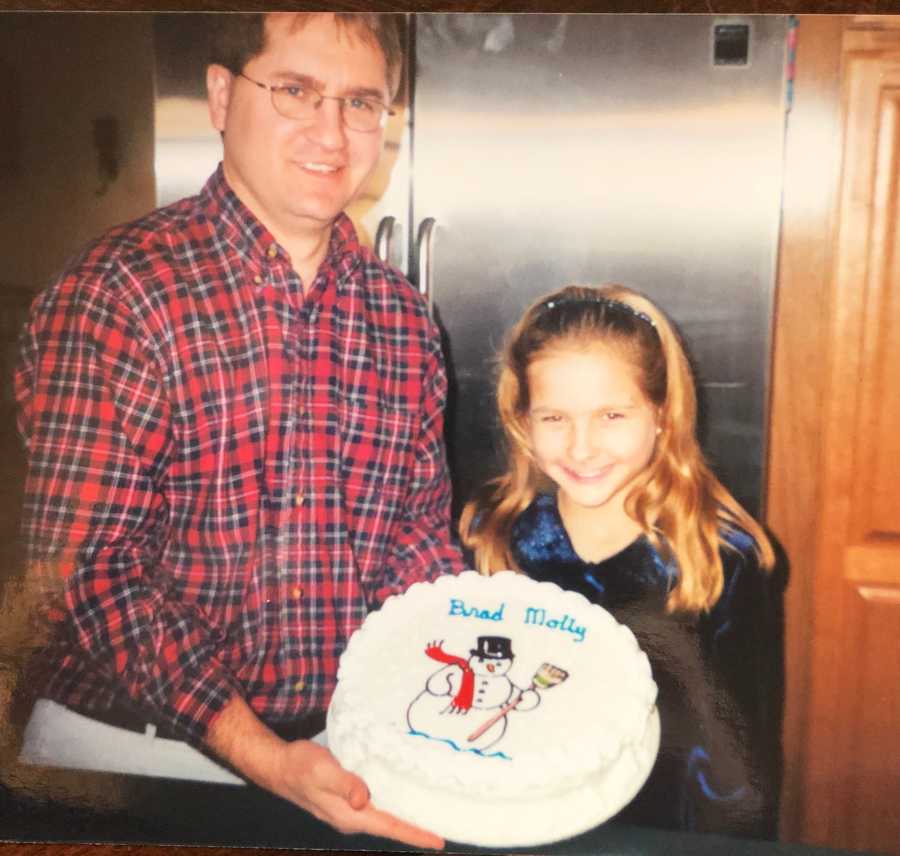 Father stands in kitchen beside daughter holding birthday cake with Frosty the Snowman on it for daughter