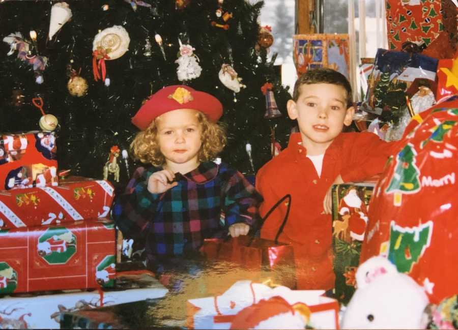 Little girl sits beside brother in front of Christmas tree surrounded by presents