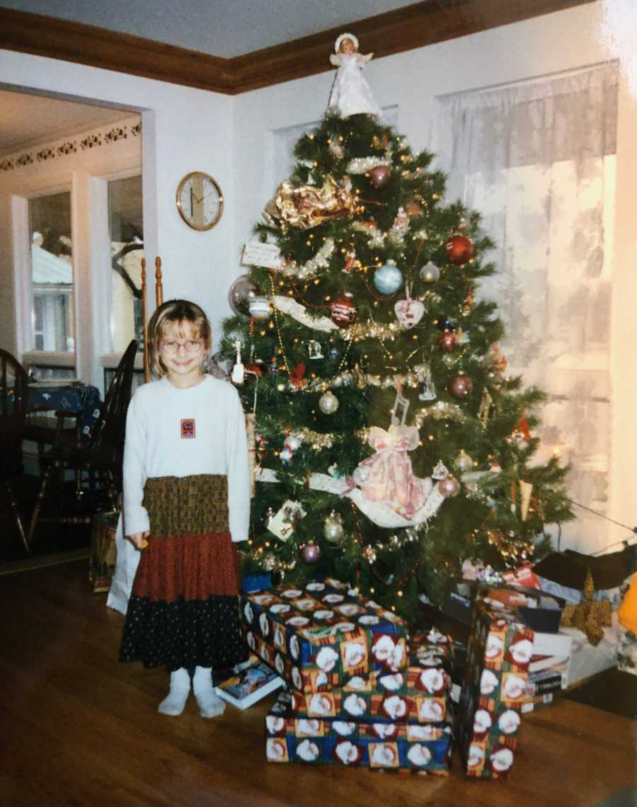 Little girl whose birthday is on Christmas Eve stands beside Christmas tree with presents under it