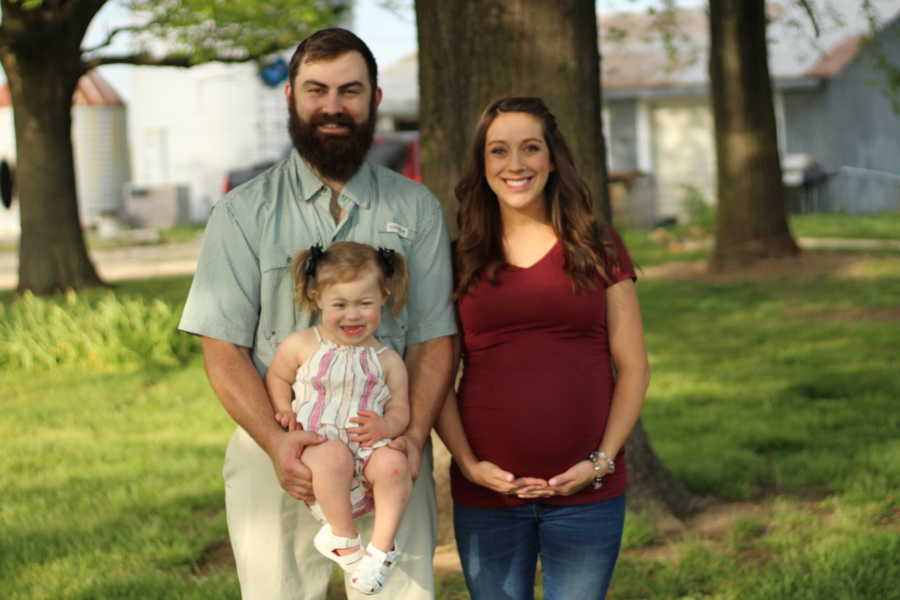 Pregnant woman stands outside holding stomach beside husband who holds daughter with down syndrome