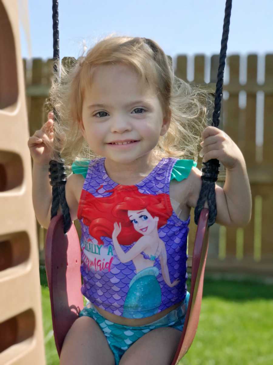 Little girl with chronic illness sits smiling on swing set wearing little mermaid swimsuit