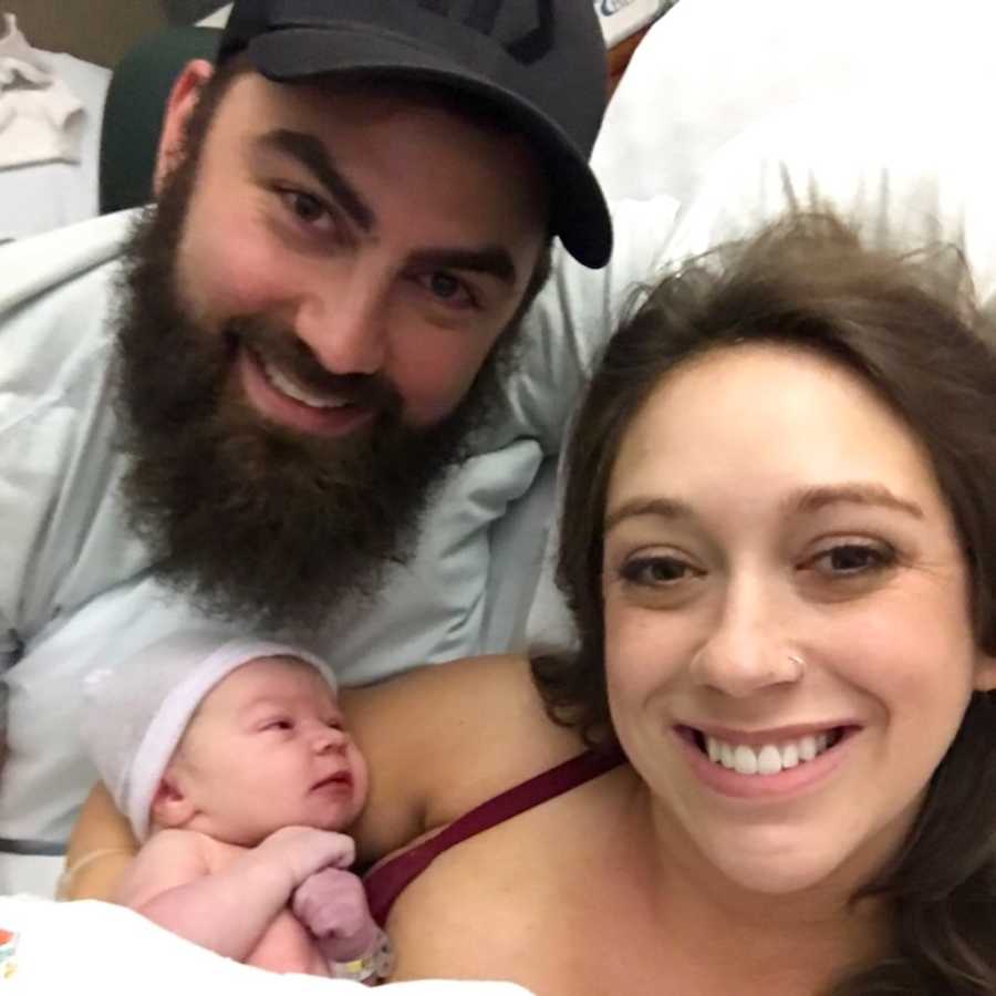 Mother smiles in hospital bed with newborn in her arm beside husband in selfie