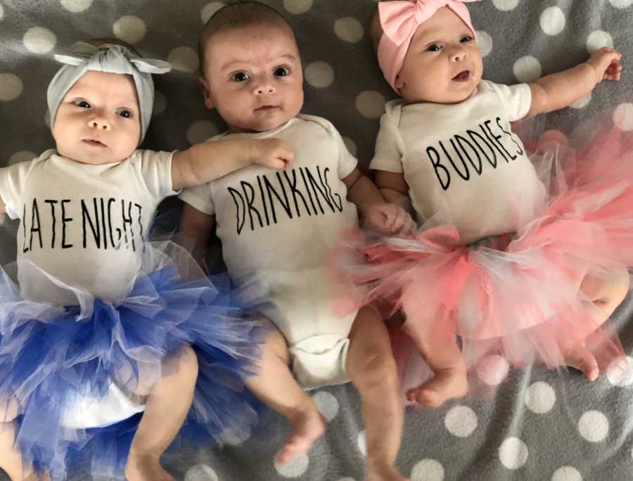 Newborn triplets lay on their backs beside each other each wearing a onesie that says, "late night" "drinking" and "buddies"