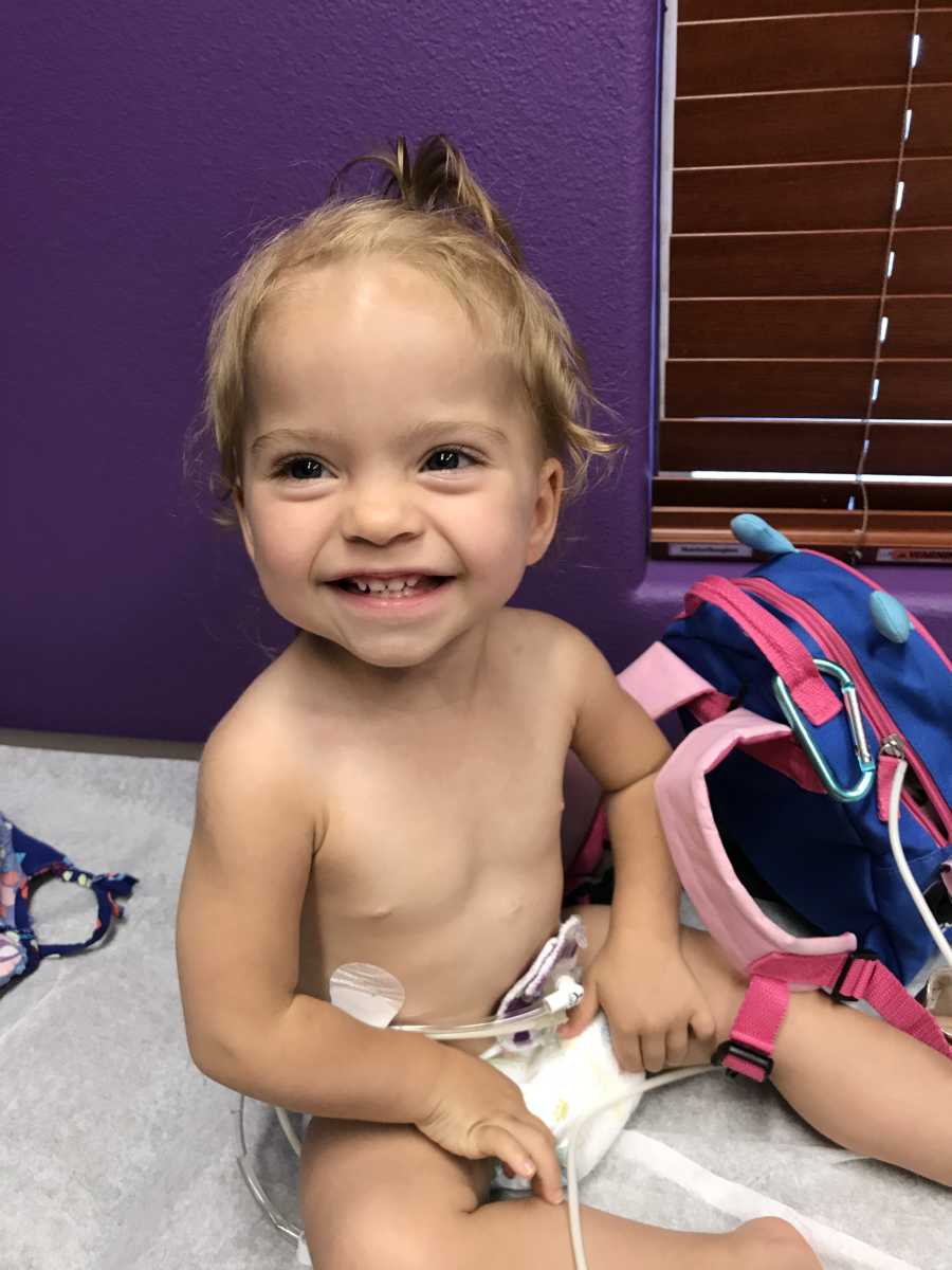 Little girl with chronic illnesses sits smiling on doctors office table only wearing diaper