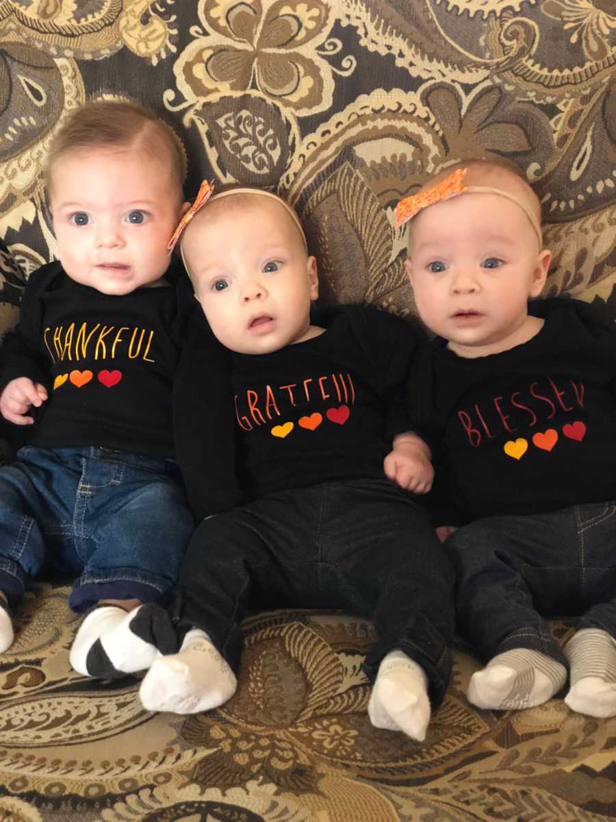 Newborn triplets sit on couch each wearing matching shirts that have "thankful" "grateful" and "blessed" written on them