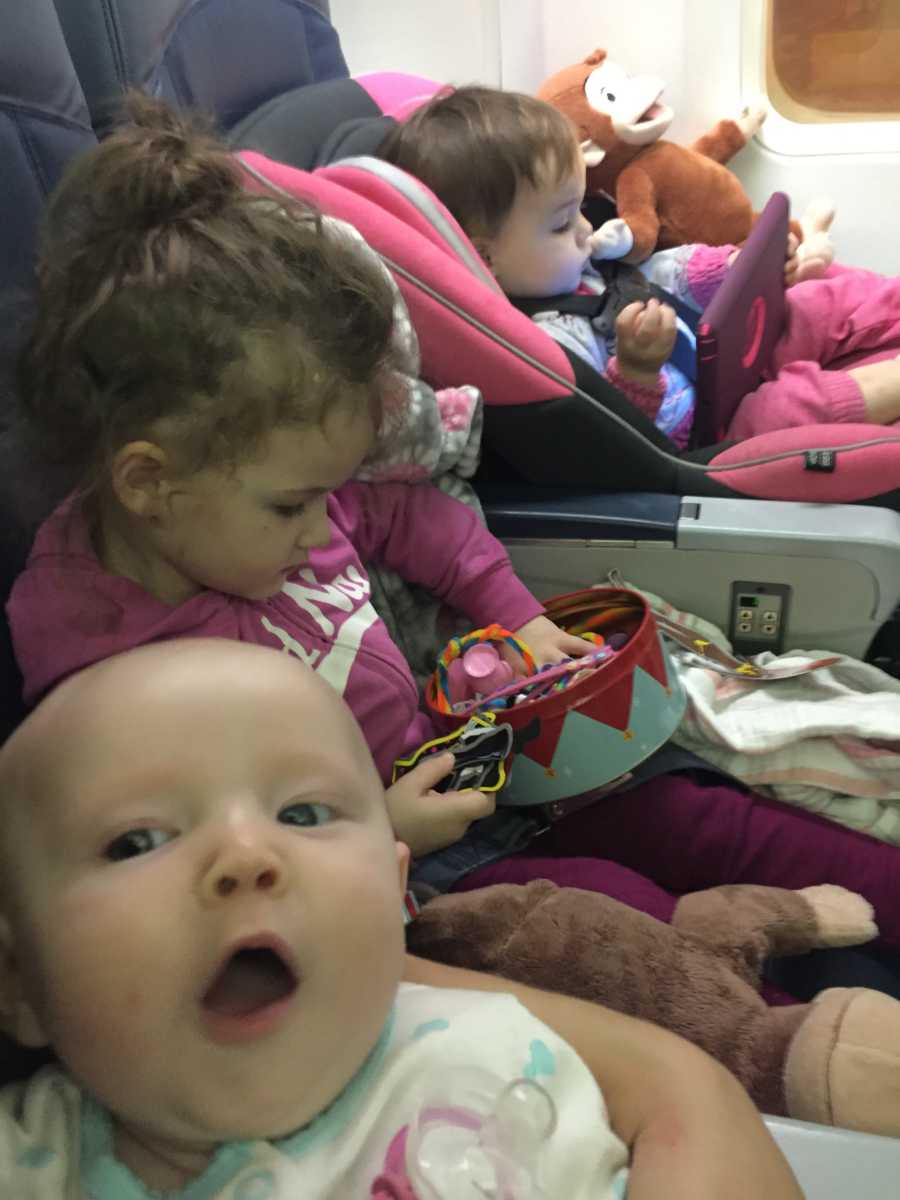 Mother takes selfie showing her baby in her arm with oldest daughter sitting beside baby in carseat in airplane row