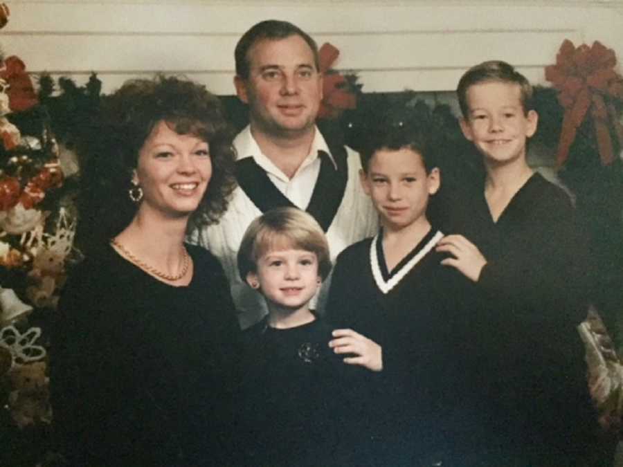 Husband and wife stand smiling with kids, one of which has since passed away, in home that is decorated for Christmas