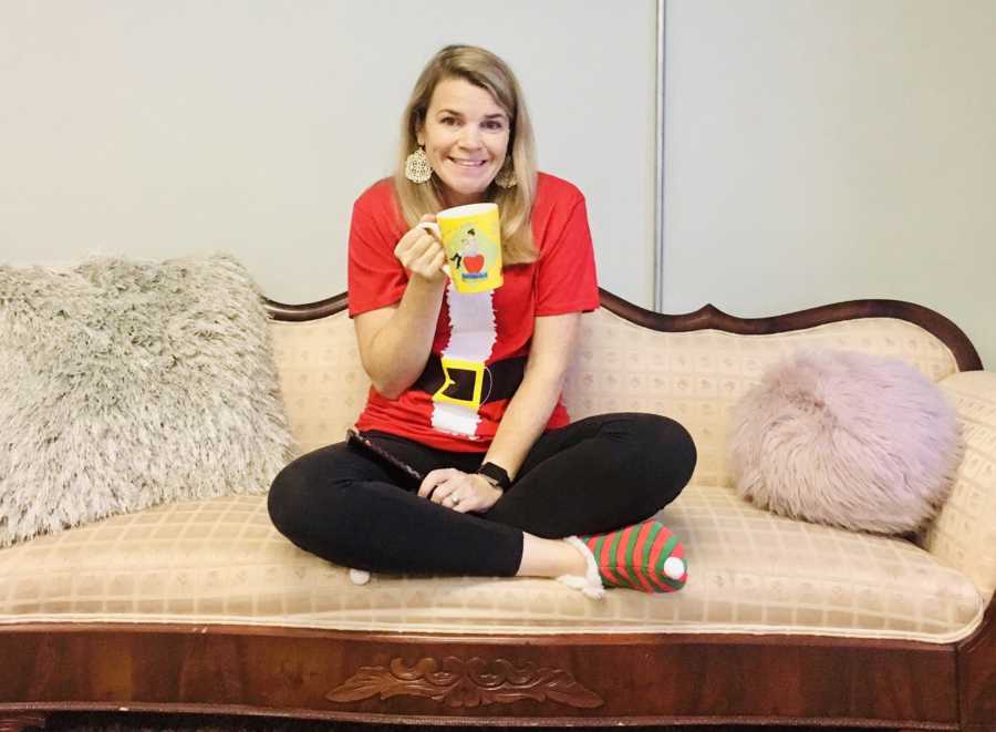 Woman who says she is addicted to Christmas movies sits on couch holding mug wearing Santa suit t-shirt and elf slippers