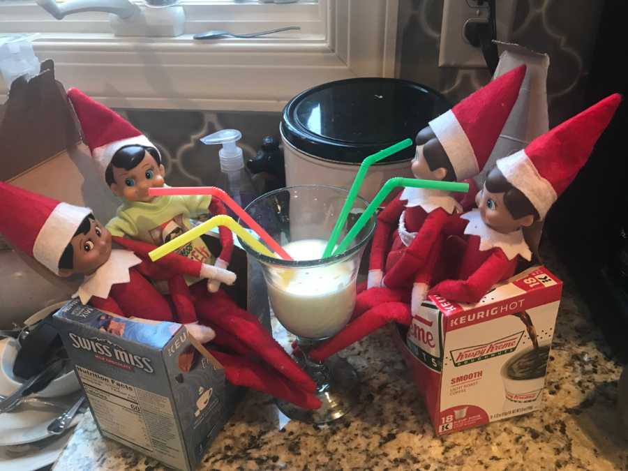 Four elves from Elf on the Shelf sit around glass of milk with four straws in it