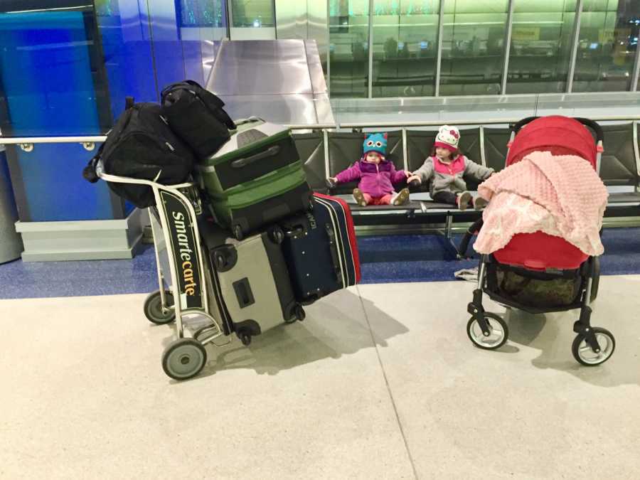 Two sister sit in chairs at airport beside stroller and luggage cart filled with luggage