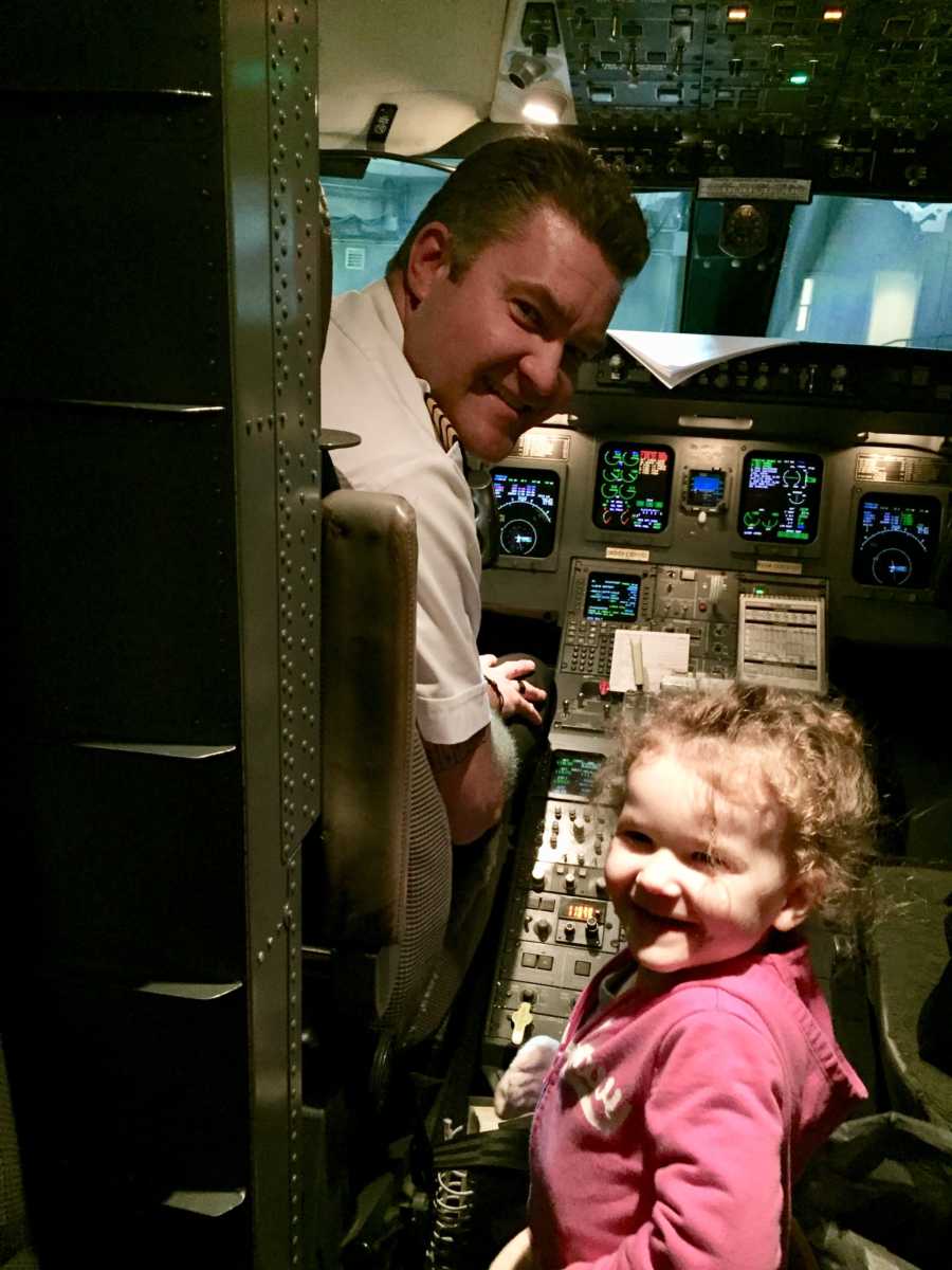 Little girl stands smiling in cock pit of plane while pilot turns around smiling