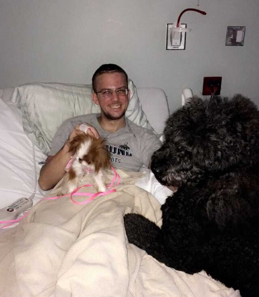 Man smiles as he lays in hospital bed with two dogs at his side