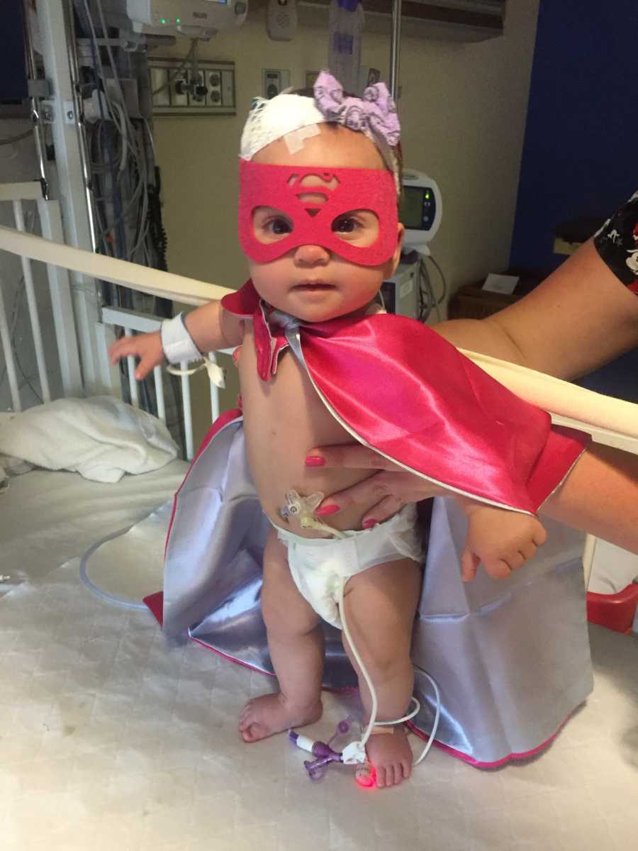 Baby girl with Celiac Disease and hyperalgesia stands in pink Superman cape and mask as she is held by nurse