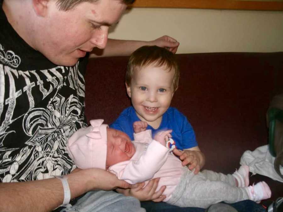 Father sits on couch beside firstborn son as they hold newborn girl