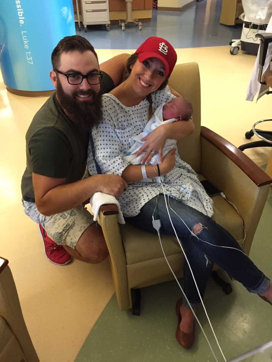 Mother sits in chair in NICU holding baby who needs open heart surgery while husband squats beside her