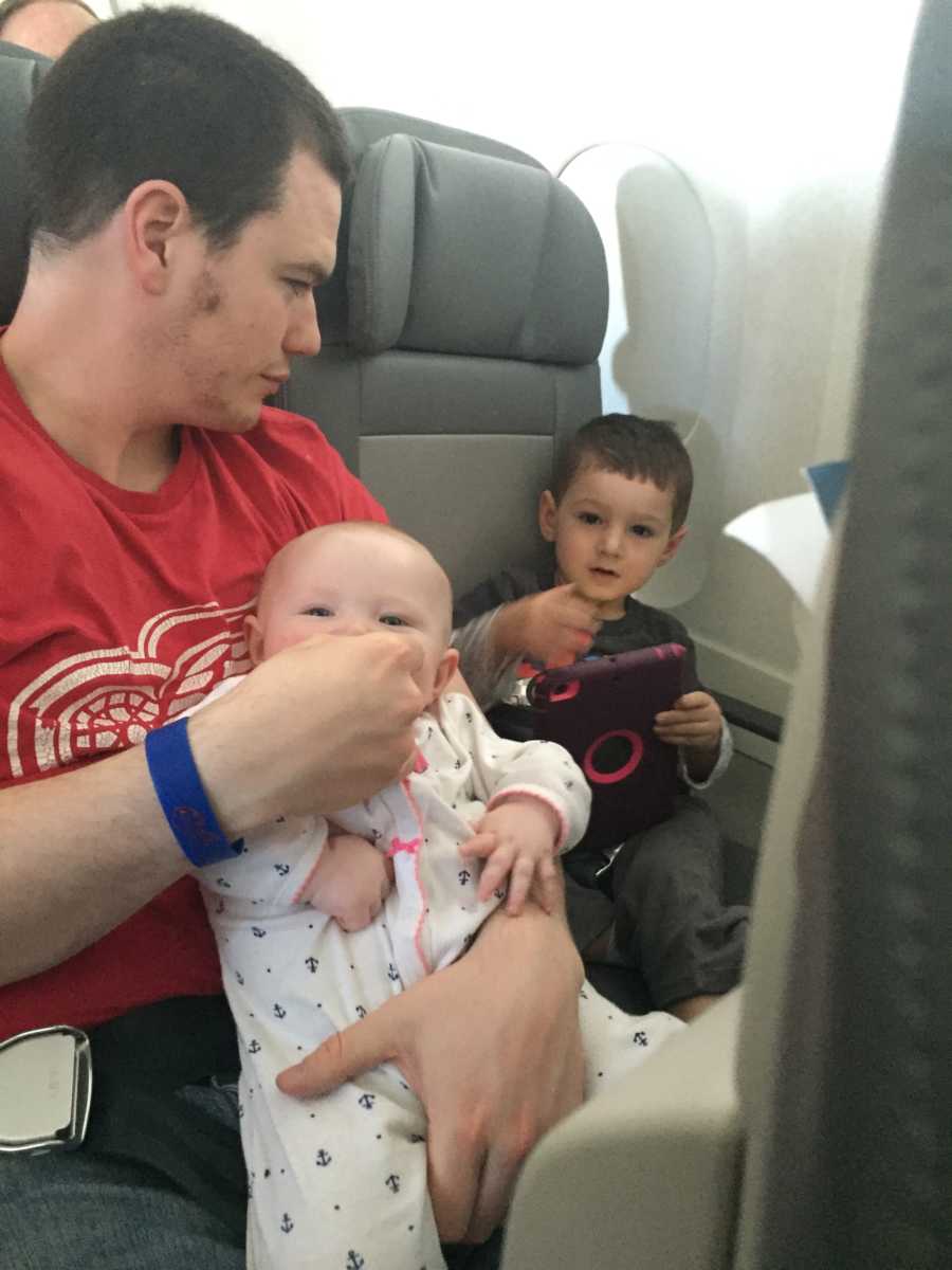 Father sits on airplane with baby in his lap beside son who sits holding iPad