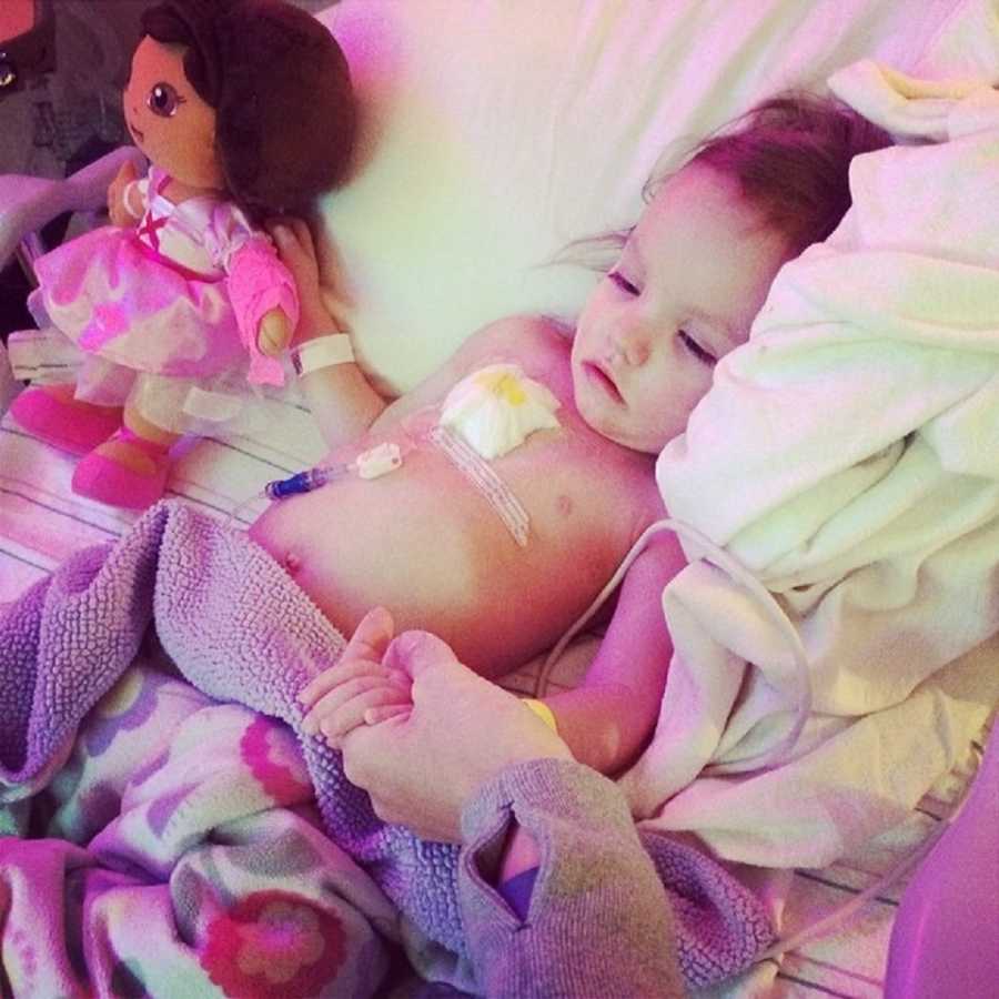 Adopted little girl with Neurofibromatosis lays in hospital bed beside toy Dora while holding parent's hand