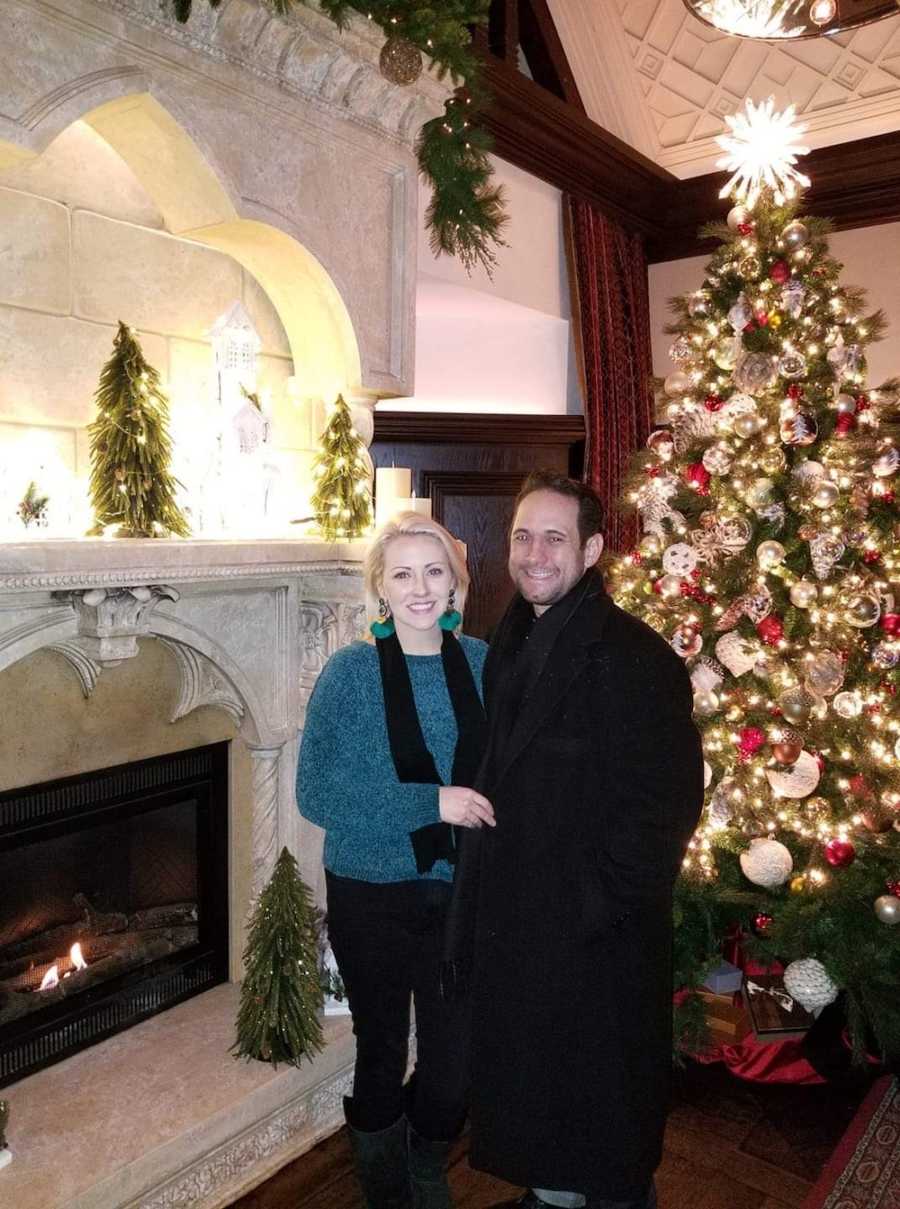 Couple smile beside fire place and Christmas tree