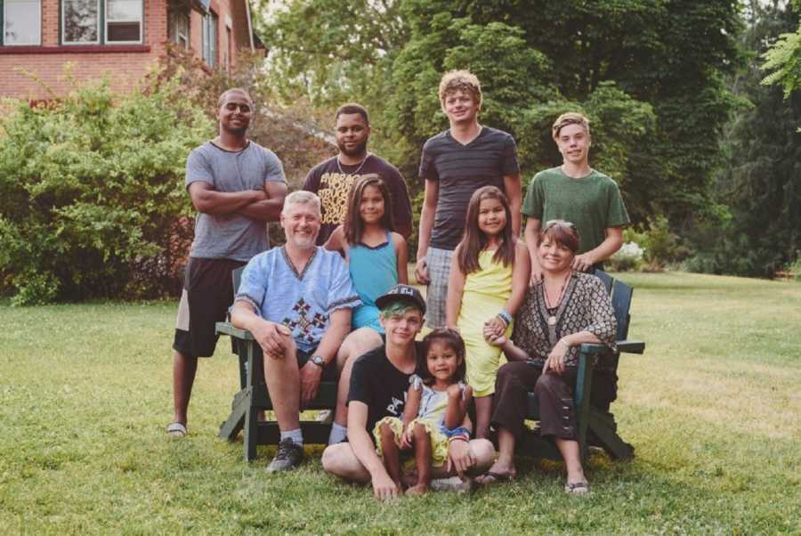 Husband and wife sit in chairs outside in their yard with their four kids and four foster children