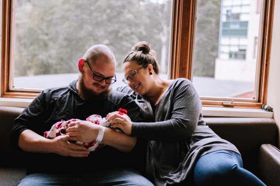 Boyfriend and girlfriend sit on couch smiling down at their baby in boyfriend's arms