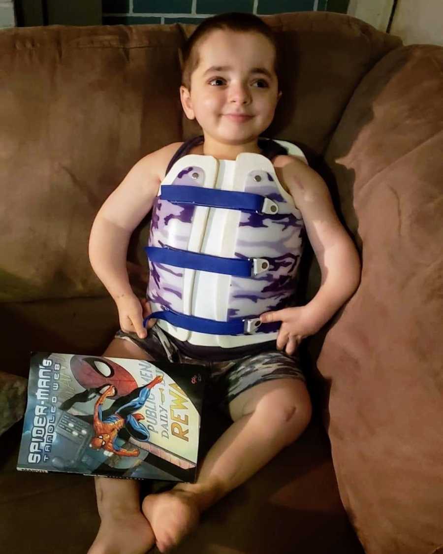 Little boy sits on couch smiling with chest brace on holding Spiderman comic book