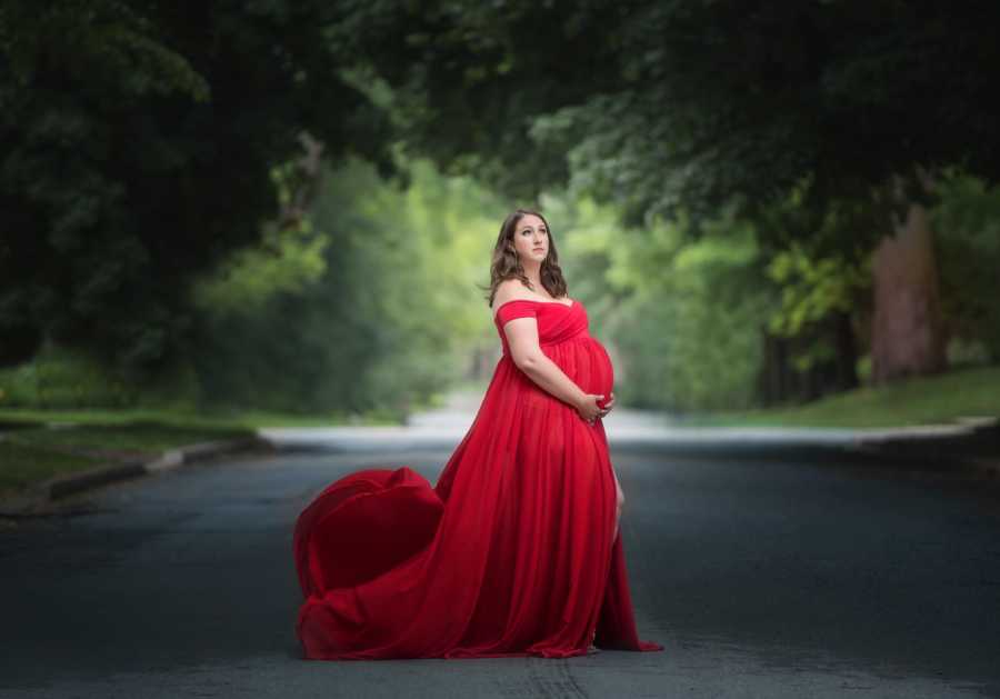 Pregnant woman stands in red gown in middle of street holding her stomach in photoshoot