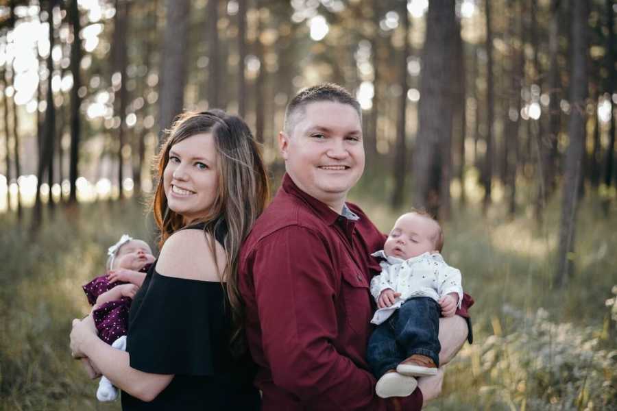 Husband and wife stand back to back in wooded area each holding twin baby