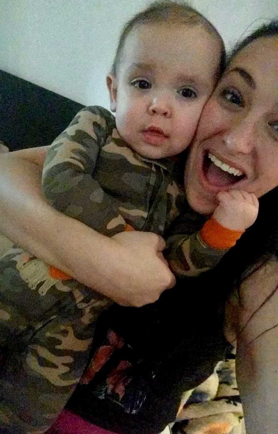 Mother smiles in selfie holding baby son in camo onesie in her arm