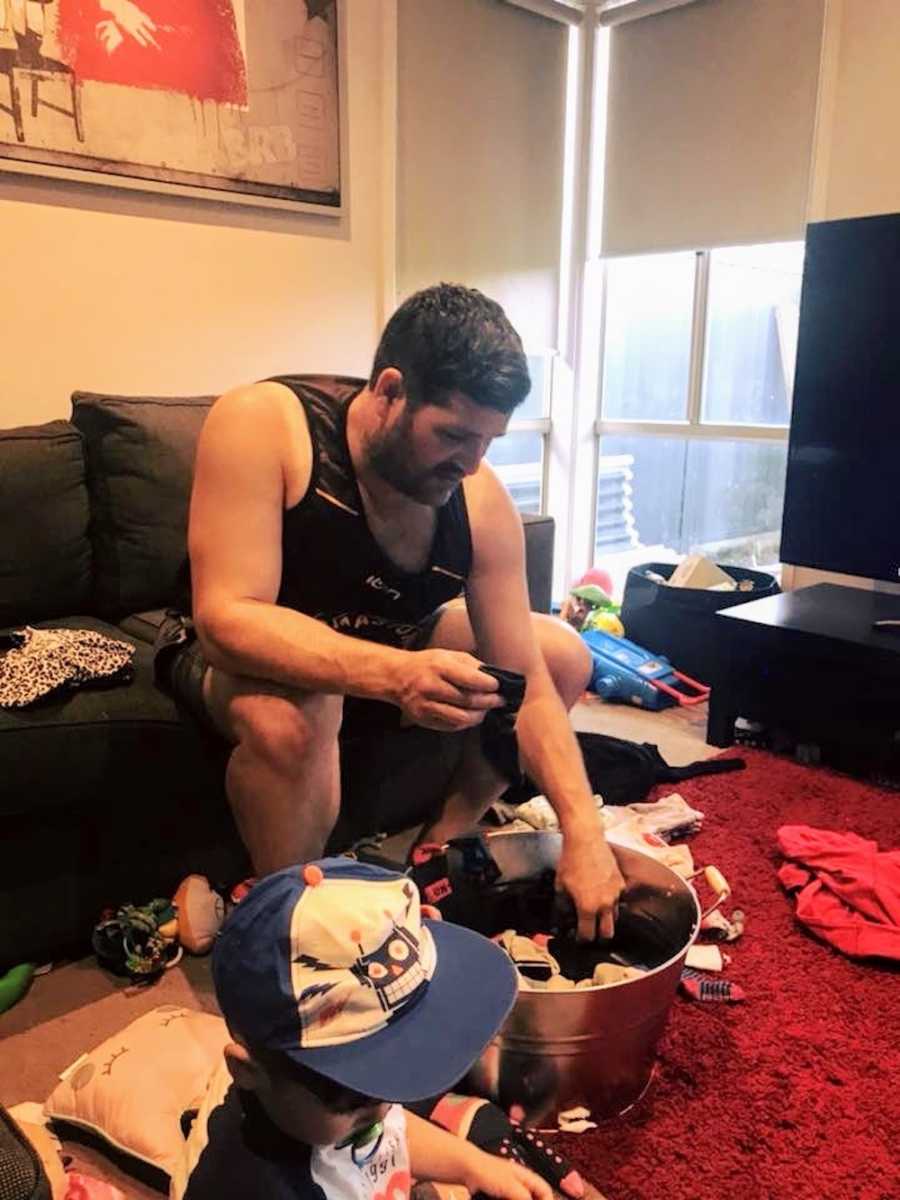 Husband sits on couch folding laundry in messy room where son sits on ground beside him