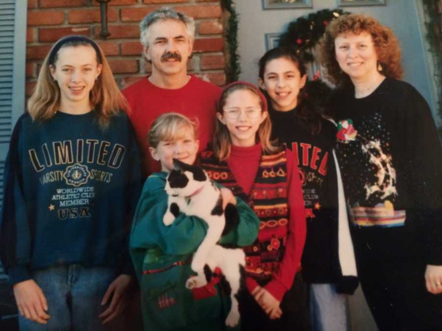 Husband and wife stand outside their front door with their four daughters and cat