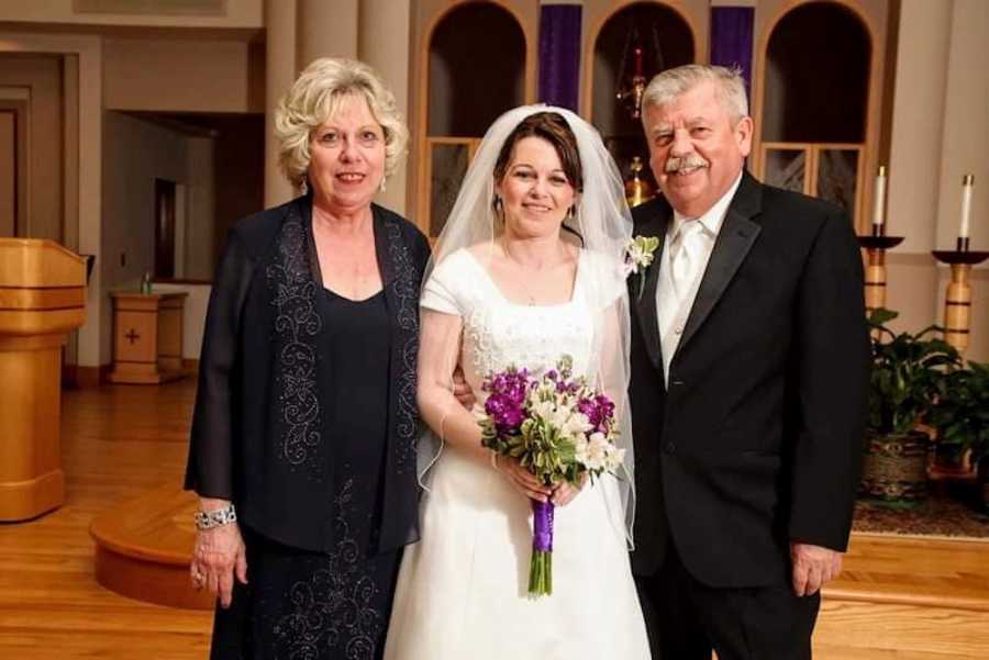 Bride stands smiling in church with adopted parents beside her