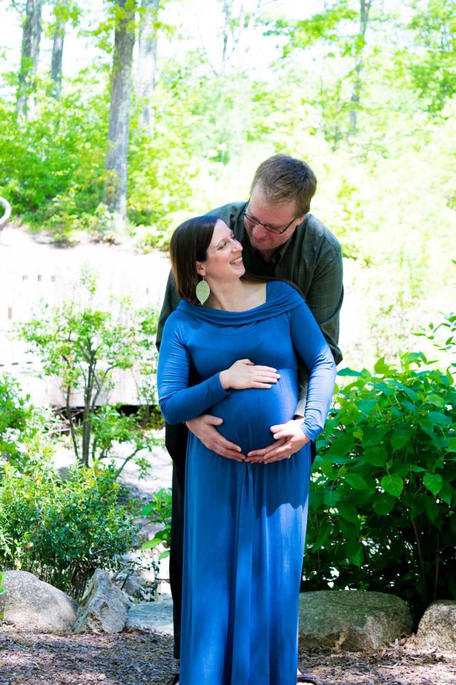 Husband stands behind pregnant wife in wooded area as they hold her stomach