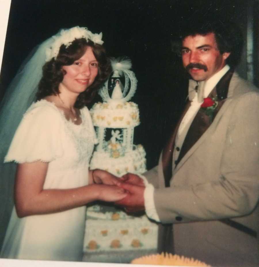 Bride and groom stand smiling holding hands with cake in background