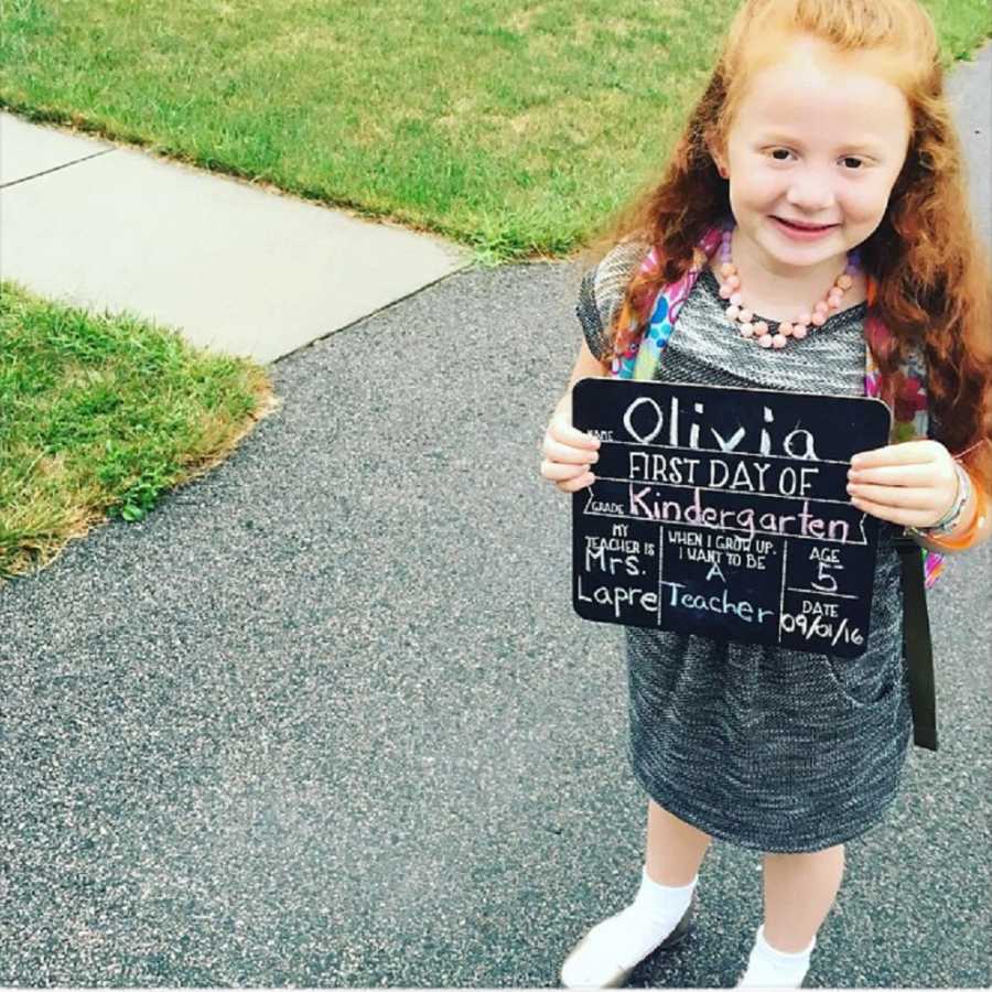 Little girl stands in driveway holding up sign saying, "First Day of Kindergarten"