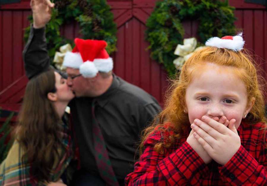 Little girl holds hands over her mouth as her father wearing Santa hat kisses her mother
