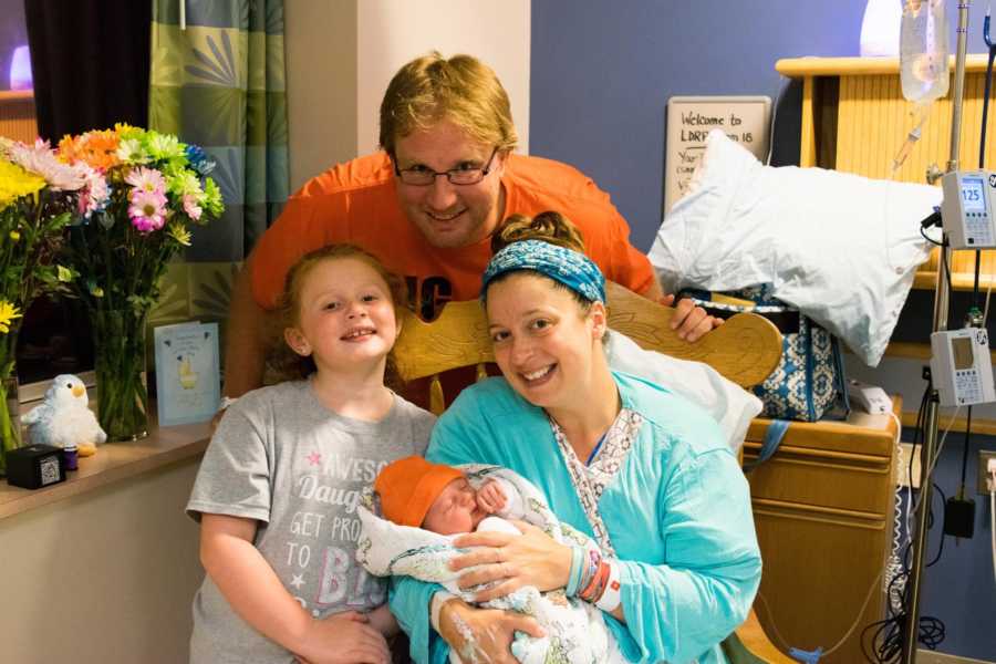 Husband stands behind wife sitting in hospital holding newborn beside their first born