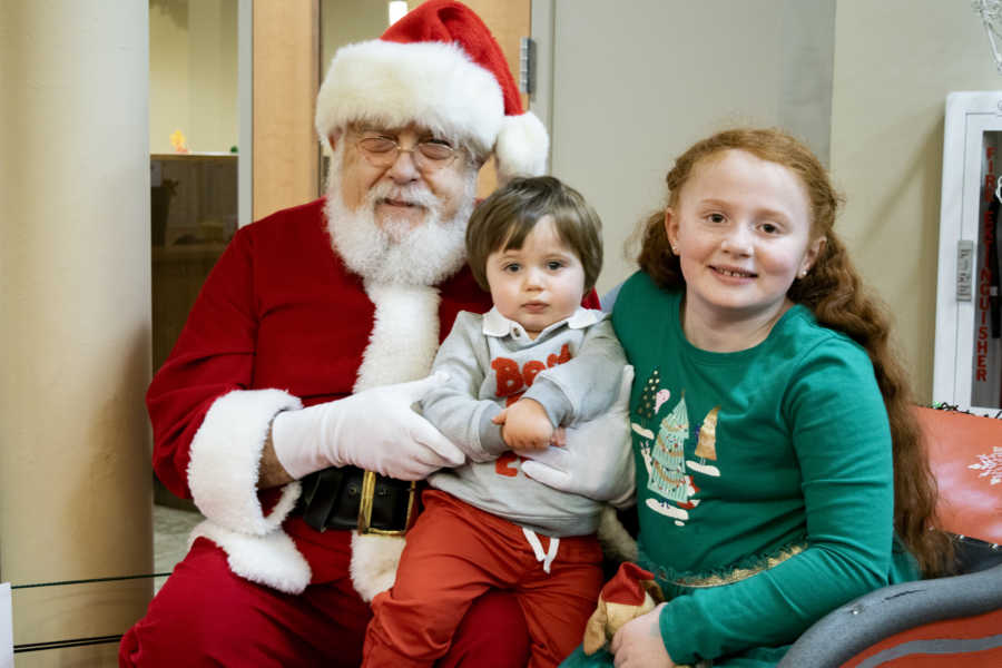 Little girl sits beside baby brother who sits on mall Santa's lap